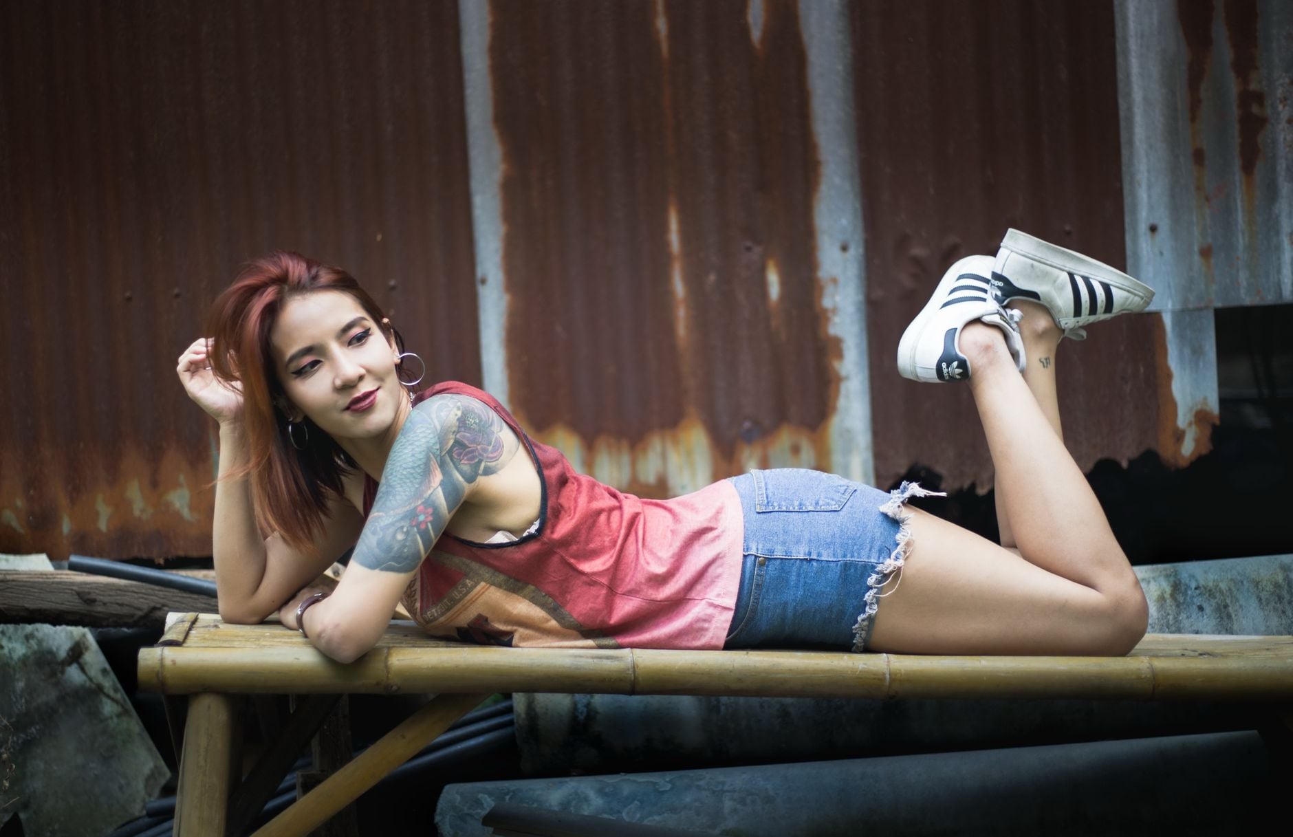A tattooed woman in casual wear poses playfully on a bench outdoors, smiling confidently.